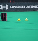 A sports field features a black and pink sports bag, two pyramids of yellow balls, and a portion of a person in dark athletic gear. The background displays a banner with the Under Armour logo.