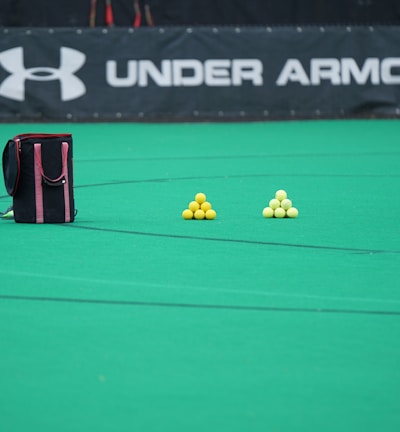 A sports field features a black and pink sports bag, two pyramids of yellow balls, and a portion of a person in dark athletic gear. The background displays a banner with the Under Armour logo.