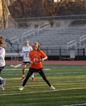 Players engaged in a competitive lacrosse match on a field, with a focus on one player in an orange jersey attempting to catch or throw a yellow ball using their lacrosse stick. Spectators and other players are visible in the background, with a stadium setting of empty bleachers.