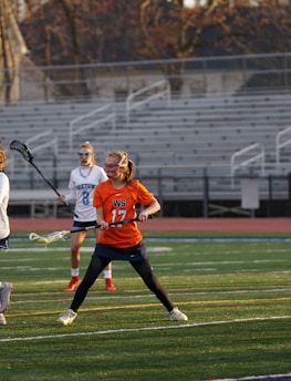 Players engaged in a competitive lacrosse match on a field, with a focus on one player in an orange jersey attempting to catch or throw a yellow ball using their lacrosse stick. Spectators and other players are visible in the background, with a stadium setting of empty bleachers.