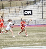 Players are engaged in a competitive lacrosse game on a grassy outdoor field. One player in a red uniform is running with the ball, pursued closely by an opponent in a white uniform. A scoreboard and stone bleachers are visible in the background.