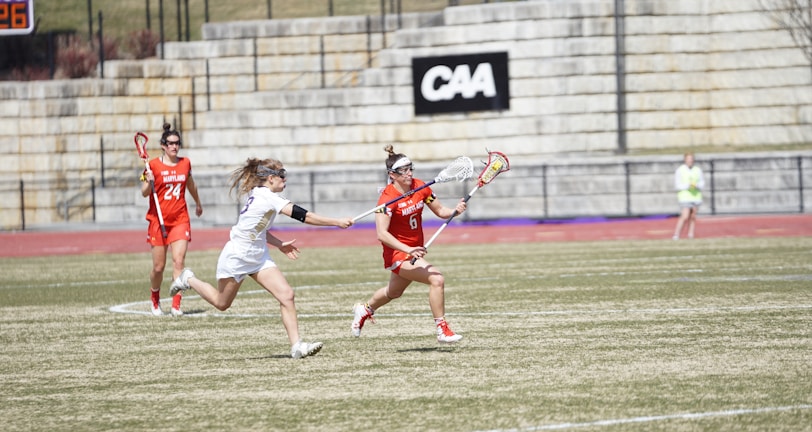 Players are engaged in a competitive lacrosse game on a grassy outdoor field. One player in a red uniform is running with the ball, pursued closely by an opponent in a white uniform. A scoreboard and stone bleachers are visible in the background.