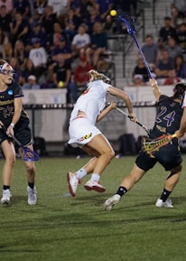 A group of athletes engaged in a competitive lacrosse game on a field. Players are wearing uniforms with numbers, and some are mid-action, reaching for the ball with their sticks. The background shows a crowd of spectators seated in stands under stadium lighting.