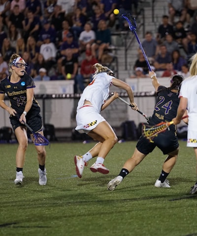 A group of athletes engaged in a competitive lacrosse game on a field. Players are wearing uniforms with numbers, and some are mid-action, reaching for the ball with their sticks. The background shows a crowd of spectators seated in stands under stadium lighting.