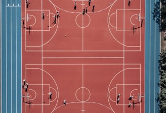 An aerial view of a large, vibrant sports complex featuring a red basketball court with multiple hoops and a surrounding blue running track. Several people are scattered across the area, engaged in various activities.