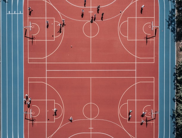 An aerial view of a large, vibrant sports complex featuring a red basketball court with multiple hoops and a surrounding blue running track. Several people are scattered across the area, engaged in various activities.