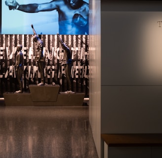 A dimly lit display features statues of athletes standing on a podium. They are positioned in front of a backdrop with the words 'LEVELING THE PLAYING FIELD' in bold letters. On the wall to the right, a softly lit quote is partially visible. The overall ambiance suggests a museum or exhibition setting.