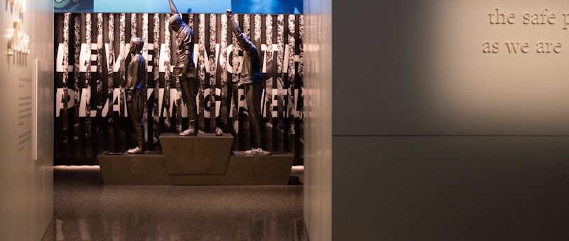 A dimly lit display features statues of athletes standing on a podium. They are positioned in front of a backdrop with the words 'LEVELING THE PLAYING FIELD' in bold letters. On the wall to the right, a softly lit quote is partially visible. The overall ambiance suggests a museum or exhibition setting.