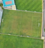 An aerial view of a sports complex featuring multiple green grass fields, including football pitches with marked goalposts. Adjacent to the fields is a blue-roofed building and a smaller area with a tennis or basketball court. Surrounding the complex, there's a road lined with trees and a dirt area with sparse vegetation.