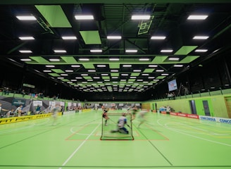 A sports hall with bright green flooring and ceiling panels. There is a game in progress with players moving quickly, suggesting a fast-paced sport like floorball or indoor hockey. Advertising banners line the walls, and a scoreboard is visible in the background.