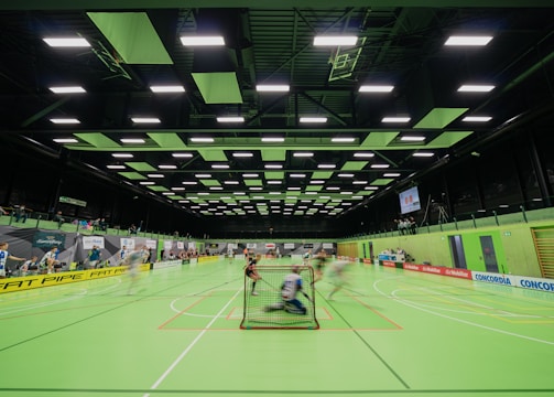 A sports hall with bright green flooring and ceiling panels. There is a game in progress with players moving quickly, suggesting a fast-paced sport like floorball or indoor hockey. Advertising banners line the walls, and a scoreboard is visible in the background.