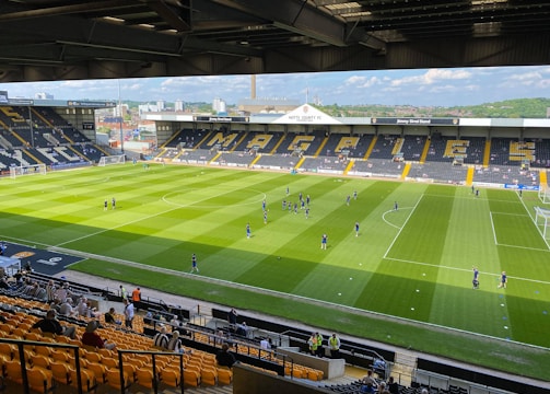 A sports stadium with a well-maintained grass field is depicted. Players are spread across the field, engaging in warm-up activities. The stands are partially filled with spectators, and the stadium is surrounded by urban buildings under a partly cloudy sky.