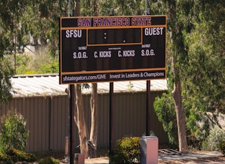 An outdoor scoreboard for San Francisco State is seen among trees and greenery. The board displays sections for shots on goal and corner kicks, with areas for both home and guest teams. It is situated near a running track with a building in the background.