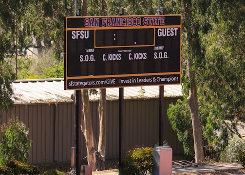 An outdoor scoreboard for San Francisco State is seen among trees and greenery. The board displays sections for shots on goal and corner kicks, with areas for both home and guest teams. It is situated near a running track with a building in the background.