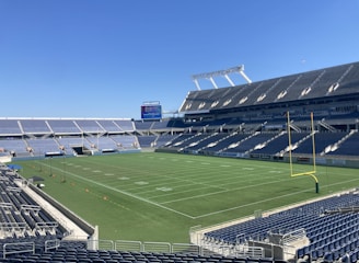 A large, empty sports stadium with blue seats surrounding a well-maintained green grass field. Bright sunlight creates clear shadows, and there are two goal posts on opposite ends of the field. A big electronic scoreboard displays some graphics, and the sky is clear and blue.